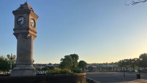 A stone tower with a clock on it surrounded by a wall near the seafront in Bangor. In the distance you can see the sails of boats. The sky is blue and is clear.
