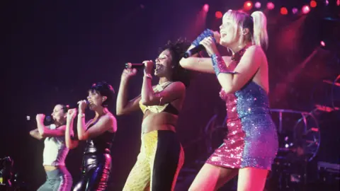 Getty Images Four singers are standing in a row singing at a concert to fans.