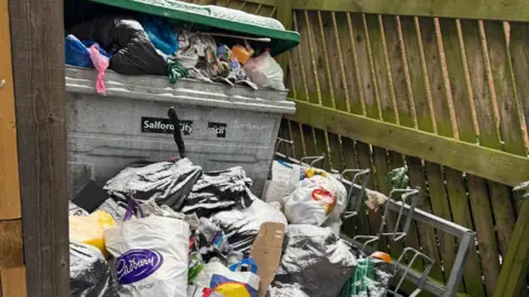Nick Barlow The inside of a bin area. The grey bin is overflowing with rubbish.
