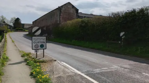 The A6 road leading into the village of Clifton of Penrith. The national speed limit signs have been turned around so the signs lead into the village instead of out. The signs read CLIFTON and there are bushes and dandelions on either side of the road.