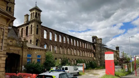 An old mill building with numerous window arches over three storeys and ornate Italianate towers at both ends.