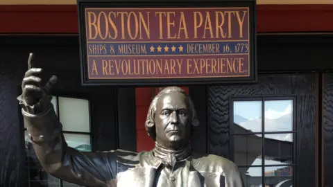 Karl Bird / BBC A red and blue sign reading "BOSTON TEA PARTY. SHIPS & MUSEUM. DECEMBER 16, 1773. A REVOLUTIONARY EXPERIENCE" in yellow capital letters. Below it is a statue of a man holding his arm up. Black doors to the museum can be seen in the background. 