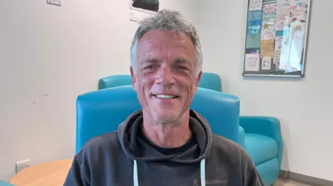 A man wearing a dark hooded sweatshirt sits in a blue chair in a hospital waiting area, with artwork and thank-you cards displayed on the walls behind him.