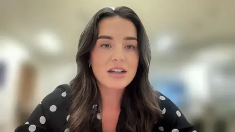 Louise Fraser, a young woman with long brown hair parted in the middle. She is facing the camera and is wearing a black top with white polka dots on it.
