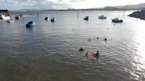 The harbour at high tide looking out towards the Bristol Channel. Boats are dotted around, while a handful of swimmers make use of the rising tide. In the distance you can see Butlins, and the hills of the Quantocks.