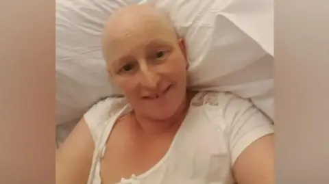 Nadeen Lister Nadeen is bald and lying in a hospital bed smiling. She is wearing a white nighty and is propped up on pillows. 
