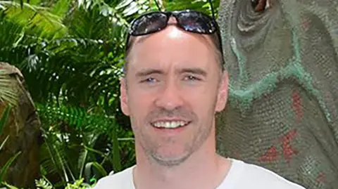 BBC Man smiling at the camera wearing a white t-shirt and sunglasses on his head. 