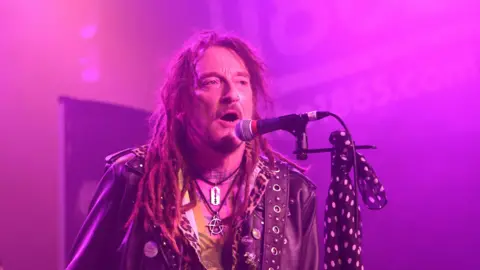 Ginger Wildheart performs with The Wildhearts at The 1865 in Southampton on 16 March 2025. He has long hair in dreadlocks, a short beard and moustache and is wearing a leather jacket and an anarchy symbol and razor blade around his neck. He has musical notes tattooed along the base of his neck and there is a blue scarf with white polka dots tied to his microphone stand. He is singing into a microphone.