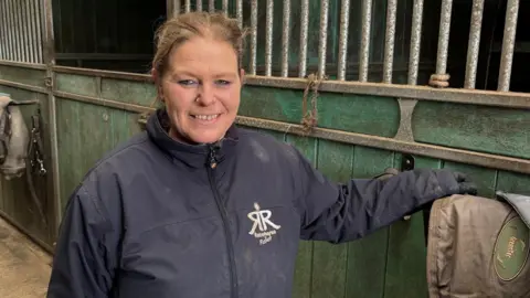 Founder of Racehorse Relief Jo Massey stood in her stable. She is looking at the camera smiling and is wearing a blue branded coat. 