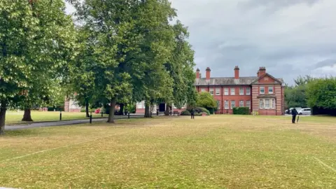An ornate building with a large field in front of it as well as a playing field and trees. There is a row of trees to the left hand side of the image, obscuring part of the building. Two people in black clothing are standing in front of the building.