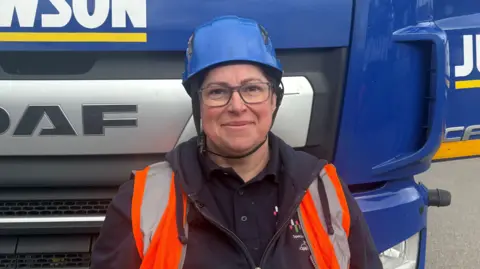 A head and shoulders photo of Nerys standing in front of a blue lorry, smiling at the camera. She is wearing a navy polo T-shirt, a zip-up hoodie and an orange hi-vis vest, which is undone. She is also wearing a blue hard hat, with a black strap under her chin, and light purple-rimmed glasses.