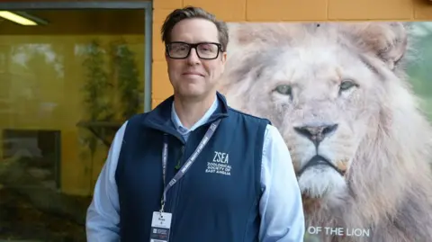 Shaun Whitmore/BBC Snipp stands in front of a lion poster. He is wearing a light blue shirt and blue zoo body warmer and a lanyard