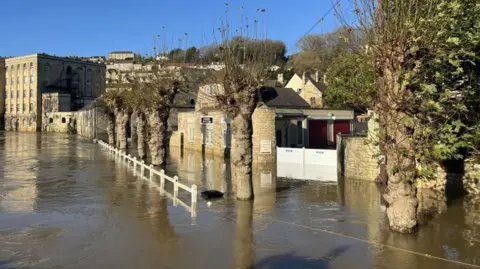 The River Avon in Bradford-on-Avon, which has broken its banks. The path next to the river is completely flooded, with the top of a white fence just visible. Water can be seen up the walls of businesses and houses. 