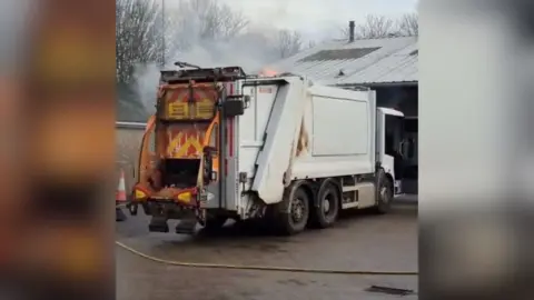 Fire and smoke emerges from the roof of a white bin lorry