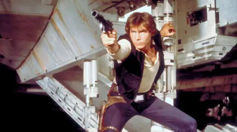 Han Solo in a screen grab from Star Wars. He has a phaser in hand about to shoot and is holding the open exit ramp to the millennium falcon 