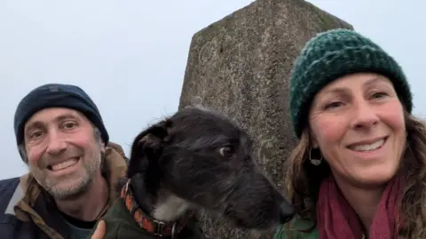 A selfie of Adam and Lou with their black dog in the middle of them after a hike. The sky is cloudy and they are sat in front of a stone. Adam is wearing a dark hat and has grey stubble, and Lou is wearing a blue woolly hat and has long brown hair. 
