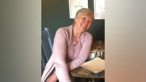 Kate Robinson, who has short blonde hair and is wearing a grey cardigan, smiles as she sits at a wooden table.