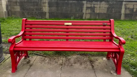 BBC/Chloe Hughes A red bench with a gold plaque on concrete slabs with grass surrounding it. There is a black bin to the left of it. Beyond the grass is a stone wall