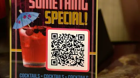 A close-up of a drinks menu on a table with a picture of a pink cocktail with a paper umbrella in it. Under the words 