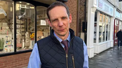 Aaron stands in a marketplace. He has a straight expression as he looks at the camera and is wearing a navy padded gilet with a gold zip over a powder blue long sleeve shirt, with a burgundy tie. Behind him is an independent homeware shop with large windows and a white painted building to the right. 