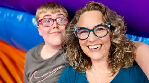 Family picture Charlotte is smiling with Adam looking to camera with his arm around her shoulder. She has curly blonde hair to her shoulders and wears horn-rimmed glasses with sparkly bits on the horns. He wears red-rimmed glasses.
