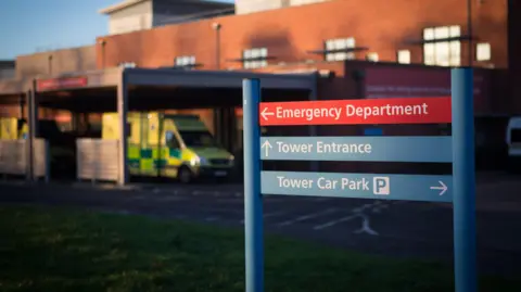 Getty Images A sign in front of a hospital, with an ambulance parked in front of it.