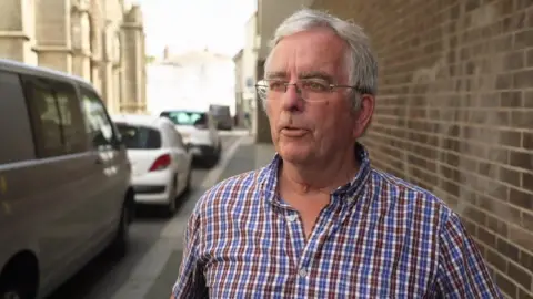A man wearing a blue, white and brown chequered top. He has glasses and short grey hair. Behind him are cars parked to the left and a brown bring wall to the right.
