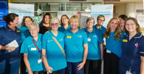 Worcestershire Acute Hospitals NHS Trust A group of 12 women stand in a hospital reception area smiling while posing for a photo. Eight are wearing bright blue polo shirts while others are in nursing uniforms and one in plain clothes, They all wear name badges.