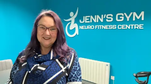 Pamela has purple curly hair and pinky black glasses. She is smiling at the camera with red lipstick on. She is wearing a dark blue flowery patterned top. The background wall behind her says 'Jenn's Gym Neuro Fitness Centre'.