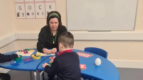 Swindon Borough Council A young boy in a navy jumper sitting at a blue table with a teacher who is wearing a black top. They are sat in a classroom with alphabet letters in the background.