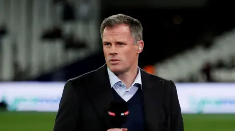 Reuters Jamie Carragher stands on a football pitch holding a microphone. He wears a light shirt, dark jumper and dark jacket. He has short greying brown hair,