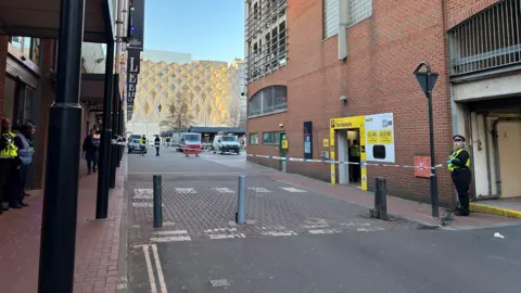 A police cordon stretches between two buildings. Officers stand on either side of the cordon. The right building features an entrance to a car park with the words "The Markets" on yellow background and a list of prices to the right.