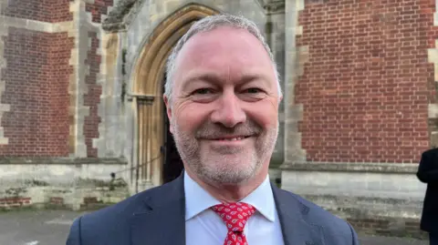 Steve Reed has short, grey hair and a beard. He is wearing a dark suit jacket, blue shirt and red and purple tie outside St Mary's Church in Slough. 