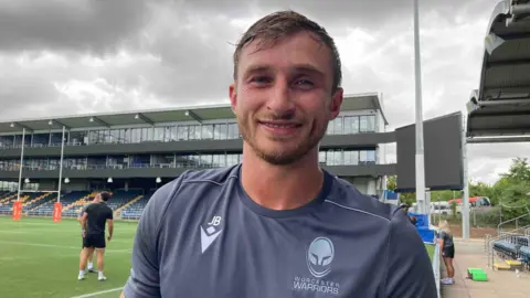 Pictured is Josh Bassett. He is smiling and has a blonde stubbly beard and wet dark blonde hair. He is wearing a grey sport t-shirt with his initials, JB, printed on the left hand side of his shirt in a light grey colour. He is stood on a rugby pitch where two people are talking in the background.