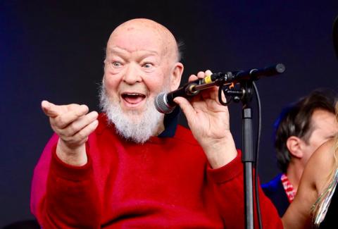 Michael Eavis performing on the Park Stage at Glastonbury Festival 2024. He is wearing a red jumper and smiling and pointing at the crowd