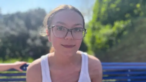 Shania, wearing a white top, looks at the camera while sitting on a blue bench.