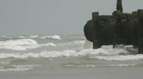 Liquid pours from a thick, black sewage overflow pipe which stands above a choppy-looking grey sea.