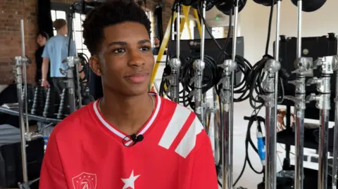 A teenager wearing a red football shirt surrounded by microphones in a studio