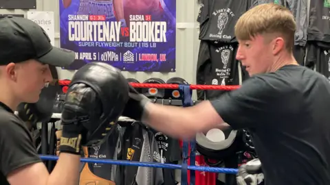 Two young men are in a boxing ring. One wears black cushioned hand pads, a black t-shirt and black cap. The other has short brown hair, a black t-shirt, black boxing gloves and is hitting the cushioned pads. In the background, there are many t-shirts and boxing posters pinned to the walls of the gym.