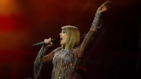 Steve Barney/BBC Taylor Swift, on stage in a sequinned outfit. She has shoulder length blonde hair and is wearing red lipstick. She is holding a microphone to her mouth, and her other hand is raised in the air.