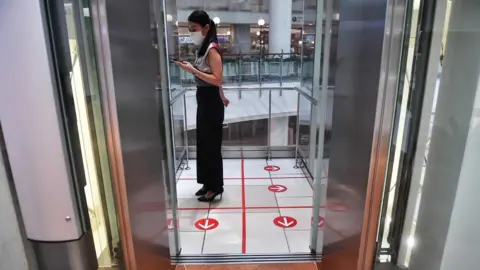 Getty Images A woman stands in an elevator with markers on the floor for social distancing measures in a shopping mall amid concerns over the spread of the COVID-19 coronavirus in Bangkok on March 20, 2020