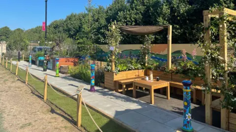 Surrey County Council Wooden bench seating in a U shape in an area the size of a standard parking space. There is also a small wooden table and green plants behind the back of the seats. Along the path are other gardens filled with green plants.
