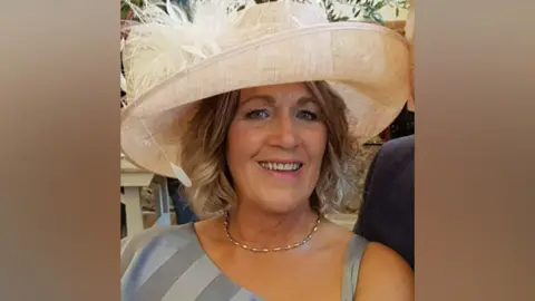 Jane Blackwell smiling at the camera. She has curly, shoulder-length blonde hair, and is wearing a large cream hat with feathers and a grey formal dress.