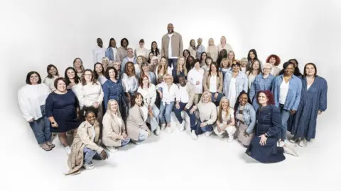 Semicircle of three rows of singers, some kneeling on the floor and others standing. They are wearing a variety of clothes, mainly blue, white and grey. 