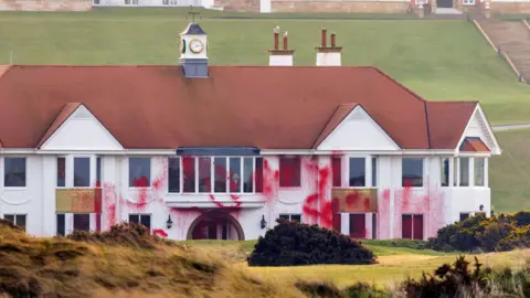 The main building at the Trump Turnberry resort in Ayrshire. The building is mainly white with a red roof. Red paint has been thrown on the side of it and partially covers some of the windows.
