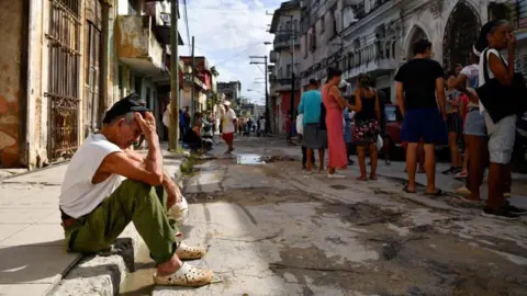 An elderly man sits on a sidewalk in the capital Havana. He looks despondent and is scratching his head, while holding a cap in his other hand. Other people mill about on the shady side of the  street.