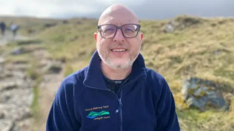 A man with glasses and a grey beard is looking at the camera. He is wearing a blue fleece with Mourne Heritage Trust written on it and is standing in front of a rocky area.