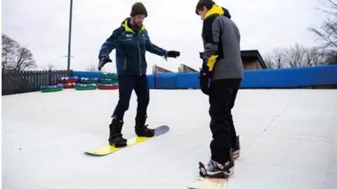 The Hill/Rossendale Leisure Trust. Two snowboarders on the Hill Ski Centre. They have warm clothes on and snow boarding boots. 