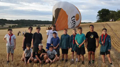 A dozen young Scout club members pose for a photo with the downed balloon, lying on its side and partially deflated, in a harvested field on a summer's evening