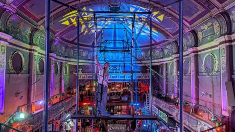 Richie Hurren A man stands on trapeze equipment, looking towards the camera inside an ornate Victorian venue with colourful lighting.